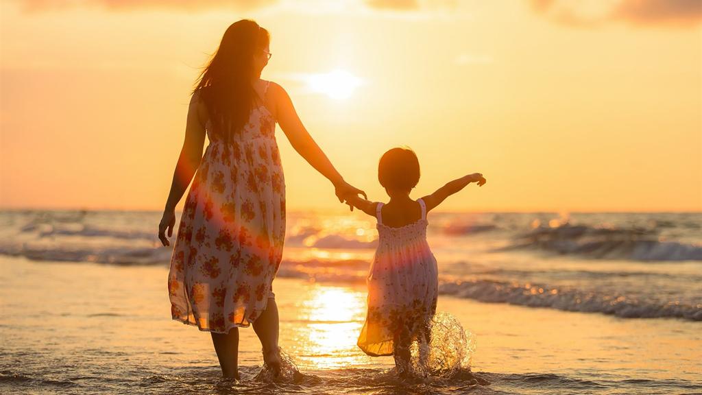 Una madre e una bambina si tengono per mano camminando nell'acqua bassa, controluce al tramonto