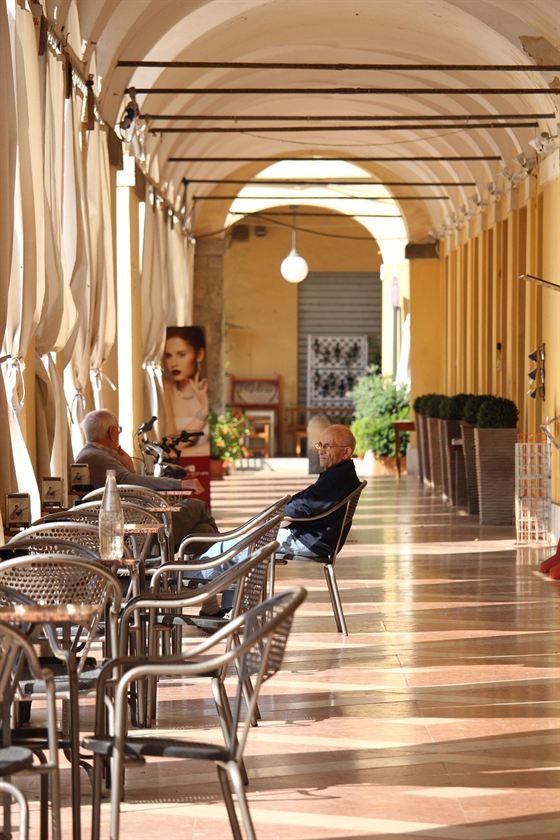 I portici del centro di Cervia, con i tavolini di un bar e due uomini seduti nella luce del pomeriggio