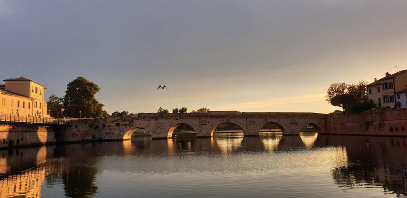 Il Ponte di Tiberio a Rimini nella luce del tramonto, le cinque arcate di pietra riflesse nell'acqua ferma e un gabbiano in volo