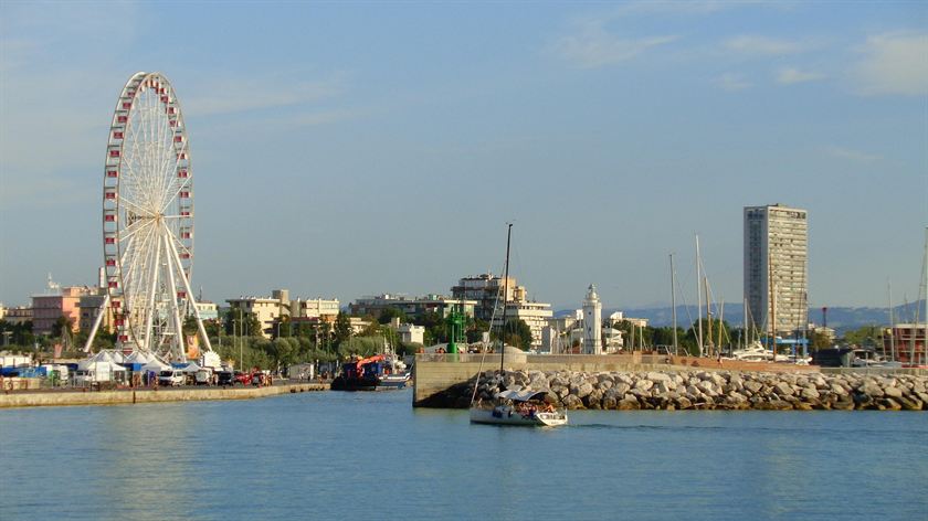 La ruota panoramica di Rimini sopra il porto, con una barca a vela che rientra fra le scogliere e il faro bianco sullo sfondo