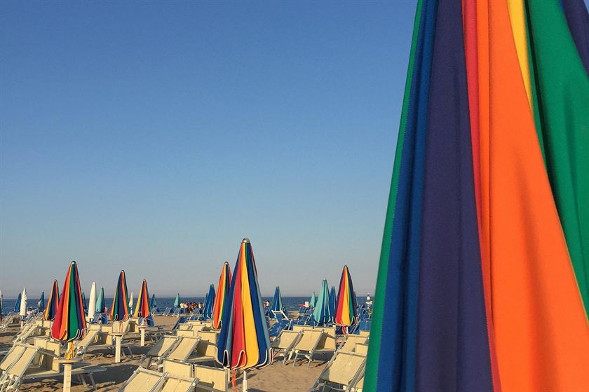 Una fila di ombrelloni ancora chiusi a spicchi colorati sulla sabbia, i lettini vuoti e il mare in fondo, sotto un cielo azzurro senza nuvole
