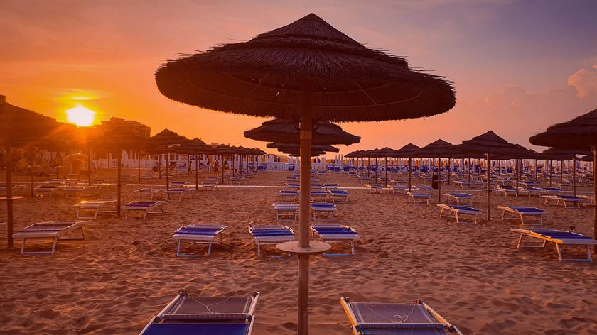 Ombrelloni di paglia aperti e lettini blu su una spiaggia, con il sole basso all'orizzonte che accende il cielo di arancione