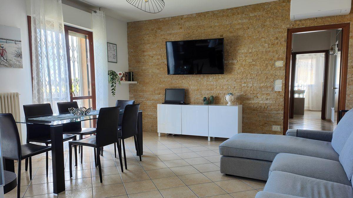 Il soggiorno di Casa Plac: il tavolo da pranzo con sei sedie, la parete in pietra chiara e il divano d'angolo